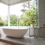 Modern bathroom interior showing free standing bath with large windows showing green trees and plants outside, with white bathtub and silver water tap, grey shower and a table with a plant.
